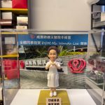 CUTEFIGUREHK 客製化醫護人員榮休公仔。一位身穿白色護士制服（肩章帶紅線）的女士，雙手在胸前比出愛心手勢，笑容親切。公仔放置在透明展示盒內，背景板印有「威爾斯親王醫院手術室 全體同事祝賀 DOM Ms. Wanda Lee 榮休」字樣及醫院外觀照片。底座前方設有印著「功成身退 樂享清閒」的祝賀牌，是送給退休護士長或醫護高管的最佳紀念禮物。