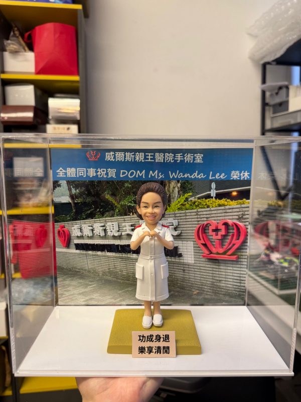 CUTEFIGUREHK 客製化醫護人員榮休公仔。一位身穿白色護士制服（肩章帶紅線）的女士，雙手在胸前比出愛心手勢，笑容親切。公仔放置在透明展示盒內，背景板印有「威爾斯親王醫院手術室 全體同事祝賀 DOM Ms. Wanda Lee 榮休」字樣及醫院外觀照片。底座前方設有印著「功成身退 樂享清閒」的祝賀牌，是送給退休護士長或醫護高管的最佳紀念禮物。