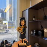 CUTEFIGUREHK 客製化女性畢業生人像公仔，生動還原了身穿黑色畢業袍、戴著四方帽的女生造型，雙手捧著一束精緻的向日葵。公仔站在黑色圓形底座上，底座設有專屬木紋牌匾，印有「Janis Chui 🎓 HKUST 2025」的字樣。這是一份專為香港科技大學（HKUST）畢業生度身訂造、極具心思的專屬畢業紀念禮物。