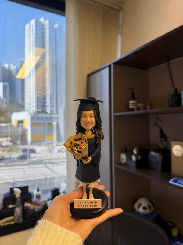 CUTEFIGUREHK 客製化女性畢業生人像公仔，生動還原了身穿黑色畢業袍、戴著四方帽的女生造型，雙手捧著一束精緻的向日葵。公仔站在黑色圓形底座上，底座設有專屬木紋牌匾，印有「Janis Chui 🎓 HKUST 2025」的字樣。這是一份專為香港科技大學（HKUST）畢業生度身訂造、極具心思的專屬畢業紀念禮物。