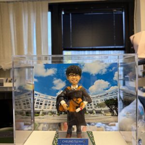 CUTEFIGUREHK 客製化男性畢業生人像公仔，生動還原了身穿黑色畢業袍、配戴橙色色帶及眼鏡的男同學造型。公仔手持畢業帽、證書及小馬毛公仔，背後配有香港科技大學（HKUST）校園風景照作為背景。底座設有藍色專屬牌匾，印有「CHEUNG Tsz Hin Happy Graduation HKUST 2025」的祝福語。整體裝在透明亞加力展示盒中，是一份極具紀念價值的大學畢業禮物。