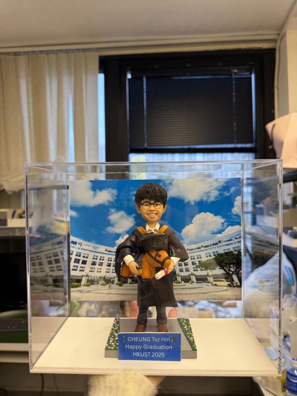 CUTEFIGUREHK 客製化男性畢業生人像公仔，生動還原了身穿黑色畢業袍、配戴橙色色帶及眼鏡的男同學造型。公仔手持畢業帽、證書及小馬毛公仔，背後配有香港科技大學（HKUST）校園風景照作為背景。底座設有藍色專屬牌匾，印有「CHEUNG Tsz Hin Happy Graduation HKUST 2025」的祝福語。整體裝在透明亞加力展示盒中，是一份極具紀念價值的大學畢業禮物。
