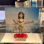 透明展示盒內的客製公仔：穿綠紅花紋洋裝的長髮人物，雙手交疊站在紅色愛心形底座上；前方有小銘牌寫著「Warranty MaMa｜Happy Retirement (2026)」。背板是天空與城市天際線，上方飛過印有「CATHAY PACIFIC」字樣的客機。畫面拍攝於室內辦公環境、可見層架與桌面物品。