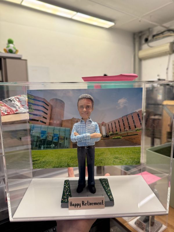 透明展示盒中的客製公仔：短灰髮角色穿淺藍格紋襯衫與深色長褲、黑鞋，雙手交叉站立；腳下為灰色步道與綠植花帶造型底座，前方銘牌寫有「Happy Retirement」。背板是紅磚與玻璃外牆的大樓草坪景色。