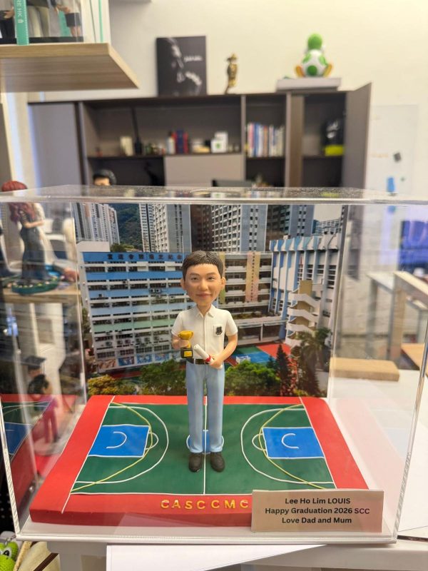 透明展示盒內的畢業主題客製公仔：短髮角色穿白色上衣與淺藍褲、黑鞋，手持金色小獎盃與捲起的證書；腳下是紅綠相間的籃球場樣式底座，上寫字母「C A S C C M C」。右前方銘牌文字為「Lee Ho Lim LOUIS｜Happy Graduation 2026 SCC｜Love Dad and Mum」。背板為高樓與校舍外觀照片。