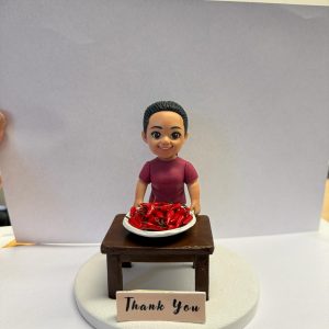 圓形白色底座上的客製公仔：紮馬尾、穿紫紅色上衣的角色站在深木色小桌後方，桌上白盤盛裝滿滿紅色辣椒；前方放置寫有「Thank You」字樣的小牌。背景以白紙遮擋，多為室內環境。
