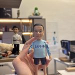 手掌托著的客製公仔：短髮角色穿淺藍色長袖運動上衣、深色短褲，赤腳站立，右手比讚；上衣正面可見深紫字樣「東方冬泳」，袖子帶有藍色圖案。背景為室內辦公環境與層架。