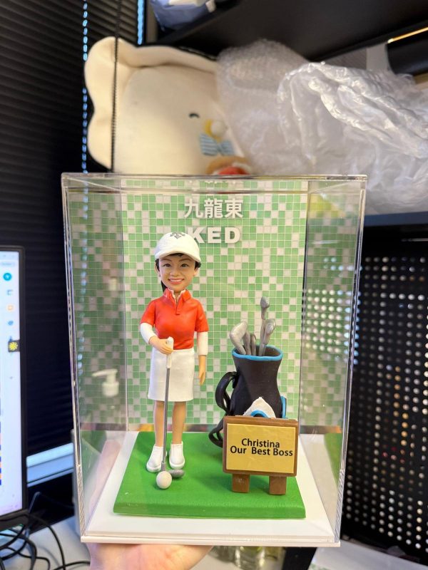 透明展示盒內的客製高爾夫主題公仔：穿白色帽、紅色高爾夫上衣與白裙的人物，手持推桿站在綠色果嶺底座上，腳邊有高爾夫球與球桿；右側放置黑藍配色的球桿袋與球桿組，前方立著小木牌，牌面印有「Christina Our Best Boss」；背板為綠色馬賽克格背景，上方可見「九龍東」與「KED」字樣。場景拍攝於室內辦公環境。