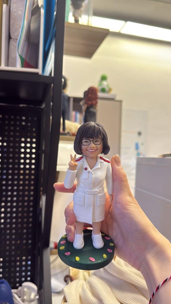 手掌托著的護理主題客製公仔：短髮戴眼鏡的人物穿全白制服，紅色肩章，右手比出勝利手勢、面帶微笑；腳踩白色球鞋，站在深綠圓形底座上，邊緣點綴粉黃小圓點。背景為室內辦公環境與層架。