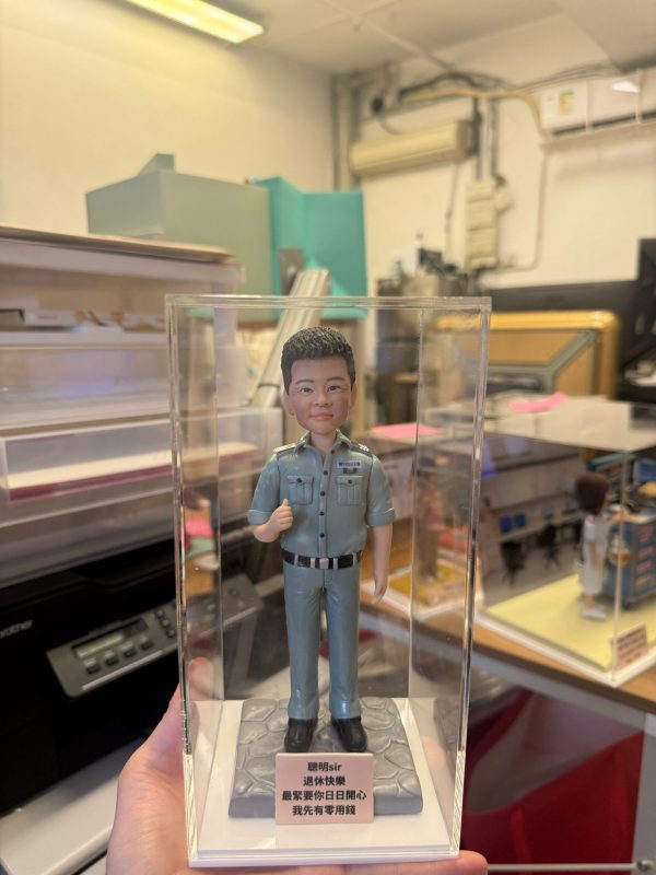 透明展示盒中的客製公仔：短髮角色穿藍灰色警務制服與黑皮帶、黑鞋，右手比讚站立；胸前有「POLICE」與階級肩章；石紋底座前粉色銘牌印有祝福語。背景為室內工作室。