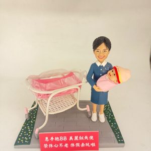 場景公仔置於灰色石板紋底座:穿藍色制服與白色鞋的角色懷抱粉色包裹的新生兒,旁邊有白粉色新生兒推床與透明床罩;底座兩側以綠色花帶點綴,前方紅色銘牌以白字寫上中文祝福語(此處不逐字轉錄),背景為淺色牆面。