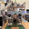 透明展示盒中的場景公仔：角色穿深色西裝、雙臂交叉站立；左右兩側各有一台附螢幕與多條管線的醫療設備推車；底板為草綠色並點綴小花片，前方銅色銘牌寫有「Thanks for being the Best Boss / Paediatric Intensive Care Unit Team / Department of Paediatrics, PWH / Year 2026」。背板為醫院外觀影像