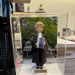 透明展示盒中的場景公仔：角色穿黑色法袍、搭配金色捲曲律師假髮與白色領巾，雙手持藍色封面的法典手冊（封面可見「Prosecution Code」字樣）；腳踩白色圓形底座，前方金色銘牌寫有「Maggie Yang / Happy retirement」。背板為綠樹與建築景色，左上角有黑白書法字樣。