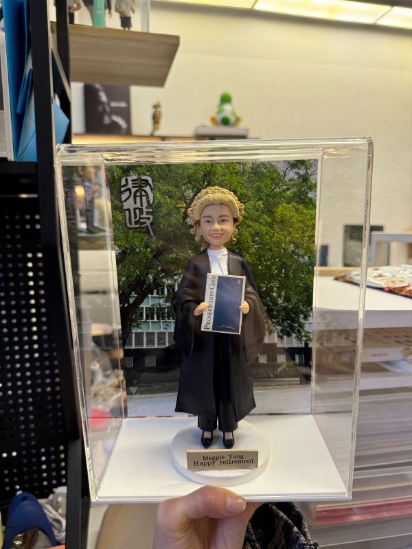 透明展示盒中的場景公仔：角色穿黑色法袍、搭配金色捲曲律師假髮與白色領巾，雙手持藍色封面的法典手冊（封面可見「Prosecution Code」字樣）；腳踩白色圓形底座，前方金色銘牌寫有「Maggie Yang / Happy retirement」。背板為綠樹與建築景色，左上角有黑白書法字樣。