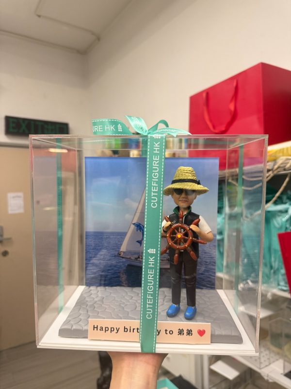 透明展示盒中的場景公仔：角色頭戴草帽與墨鏡，穿深色背心與藍色鞋，雙手握著棕色船舵；背板為藍色海面與白色帆船圖像；底座為灰色石板紋，前方長條銘牌寫有「Happy birthday to 弟弟」