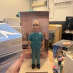 手掌托起的場景公仔：角色穿藍綠色短袖醫護制服與同色長褲、配戴眼鏡與黑色鞋，髮髻紮起；站在米色砂感長方形底座上，背板為室內科室或藥局場景。前方金色銘牌印有多行中文：「全哥！榮休之喜 多謝勞苦功高 律敦治及鄧肇堅醫院藥劑部」。