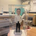 透明展示盒中的場景公仔：角色著白色西裝外套與白襯衫、搭配黑長褲與黑鞋；站在灰色圓形底座上，前方白色銘牌印有「Chairman / Chae Soo Chan」文字。背景為室內工作室與收納設備。