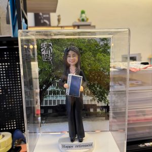 透明展示盒中的場景公仔:身著黑色西裝與黑鞋的角色手持深藍色文件手冊,站在圓形灰色底座上;前方白色小銘牌寫有「Happy retirement」;背板為綠樹與現代建築外牆,左上角有直排漢字。