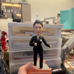 手持展示的一尊武術主題公仔:角色穿黑色道服與黑帶,胸前貼有小型國旗貼章,雙手呈出拳姿,腳下為灰色圓形底座;前方橘色銘牌印有英文字職稱與姓名(此處不逐字轉錄),背景為工作桌與收納抽屜。