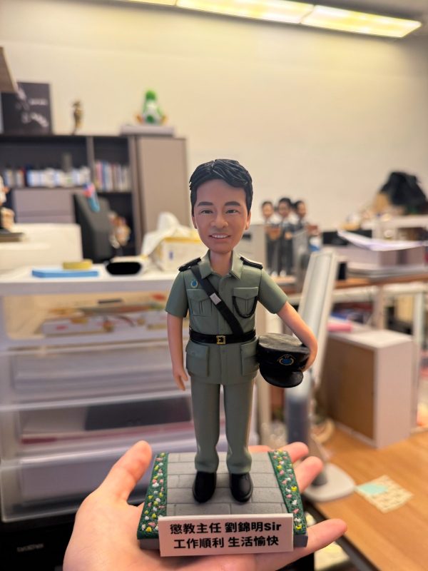 手持展示的一尊制服主題公仔：角色穿淺綠色制服與黑色皮帶，肩章細節可見，右手持深色軍帽站在灰色石板紋底座上；底座邊緣有綠色與黃色小花裝飾，前方白色銘牌以中文寫有職稱與祝福語（不逐字轉錄）；背景為辦公室與收納抽屜。