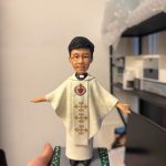 手掌托起的場景公仔：角色穿白色披肩式禮服，胸前有紅色聖心圖樣與垂直金色刺繡花紋；雙臂微展、腳穿黑鞋；站在灰色石板紋底座上，兩側綠色邊條點綴小花。前方銅色銘牌寫有「Ordination blessings for Fr. Benedict Shum 13 • 6 • 2026」。背景為室內書架與收納櫃。
