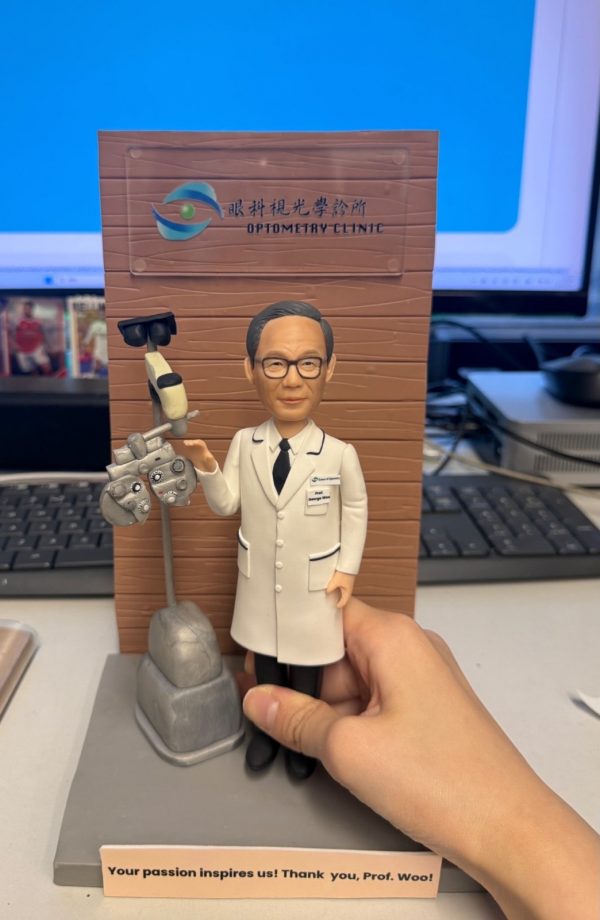 場景公仔立於灰色底座：角色戴黑框眼鏡、穿白色醫師袍與黑色領帶，旁邊有驗光儀器（phoropter）與檢查燈裝置；背後為木紋直立背板，印有眼科/驗光診所的中英文標識；前方桃色銘牌以英文寫有感謝語（此處不逐字轉錄）；背景可見鍵盤、螢幕與桌面，右側有手扶住底座。