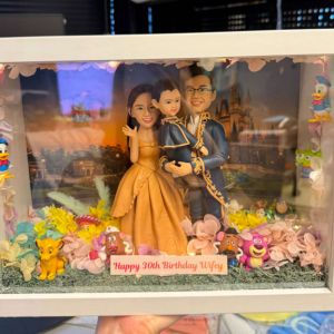 白色相框展示盒中的場景公仔:一對穿晚禮服的成人與一名幼童站在中央,背景是夕陽下的城堡畫面;腳邊鋪有草地與粉彩花瓣,四周擺放多個卡通風小擺件;底部銘牌寫有「Happy 30th Birthday Wifey」。