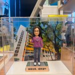 透明展示盒中的場景公仔：角色戴黑框眼鏡、穿紫色針織上衣（胸前有月桂環圖樣）、藍色牛仔褲與白鞋；站在灰色石板紋底座上，兩側綠邊點綴小花。前方淺色銘牌寫有「春風化雨，桃李滿門」。背板為校園大樓與綠樹景色，左側立柱可見多行中文校名文字。