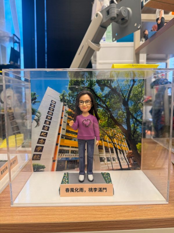 透明展示盒中的場景公仔：角色戴黑框眼鏡、穿紫色針織上衣（胸前有月桂環圖樣）、藍色牛仔褲與白鞋；站在灰色石板紋底座上，兩側綠邊點綴小花。前方淺色銘牌寫有「春風化雨，桃李滿門」。背板為校園大樓與綠樹景色，左側立柱可見多行中文校名文字。