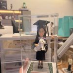 透明展示盒中的場景公仔：角色穿黑色學袍配淡紫領緣、戴學士帽與流蘇，懷抱一隻白色小型犬；站在灰色石板紋底座上，兩側綠邊點綴小花。前方淺色銘牌寫有「Congratulations」。背景為室內工作桌與收納櫃。