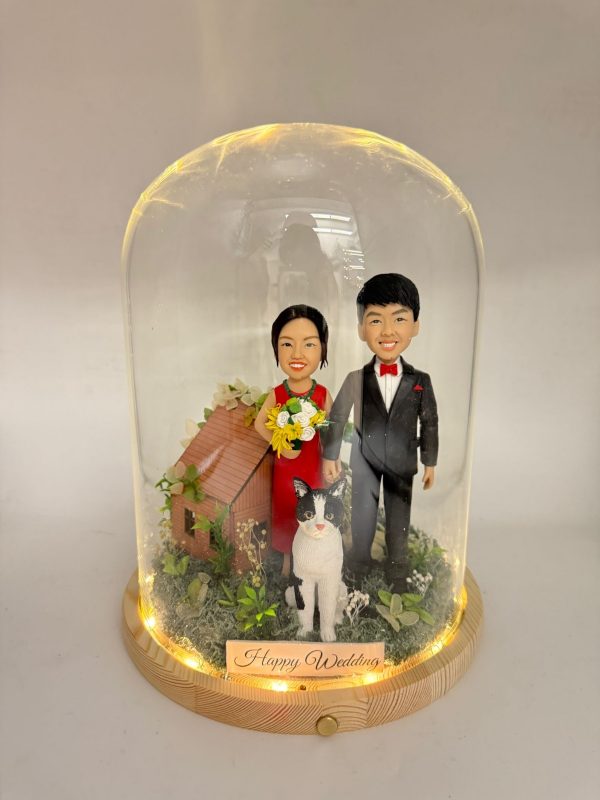 玻璃罩中的婚禮場景公仔：新娘穿紅禮服手捧黃白花束，新郎著黑色西裝配紅色領結與口袋巾；前方有一隻黑白相間的貓站立，其後為小木屋造型與綠植苔面。木質圓形底座周圍繞有暖色燈串，前置金色銘牌寫有「Happy Wedding」。