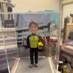 透明展示盒中的場景公仔：角色穿螢光黃綠色上衣搭配黑色長袖與長褲，腳穿運動鞋，右臂夾著黃色安全帽；站在灰色六角石板紋底座上。背板是一棟白色多層建築，屋前廣場停有紅色消防車並掛有旗幟。