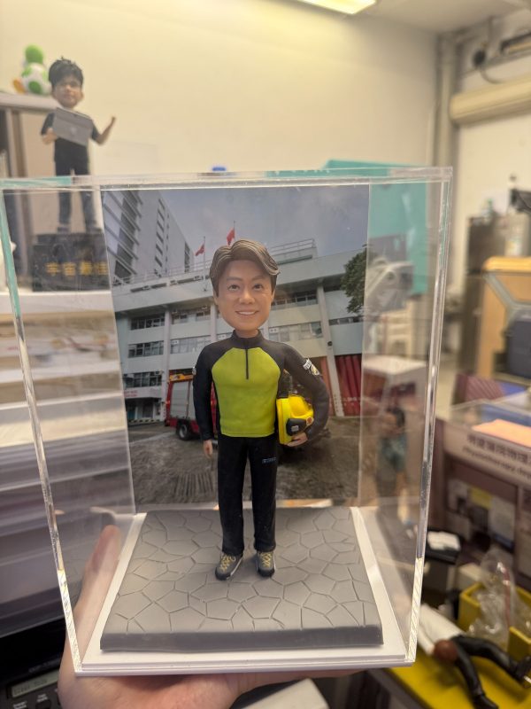 透明展示盒中的場景公仔：角色穿螢光黃綠色上衣搭配黑色長袖與長褲，腳穿運動鞋，右臂夾著黃色安全帽；站在灰色六角石板紋底座上。背板是一棟白色多層建築，屋前廣場停有紅色消防車並掛有旗幟。