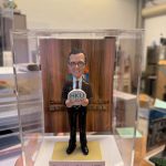 透明展示盒中的場景公仔：角色穿黑色西裝與領帶、戴圓框眼鏡，雙手捧圓形標牌，上有「HKU LAW」字樣；足下為淺金色方形底座。前方淺色銘牌寫有「Edmond Happy Retirement 2026」。背板為直紋木質背景並見英文字樣裝飾。