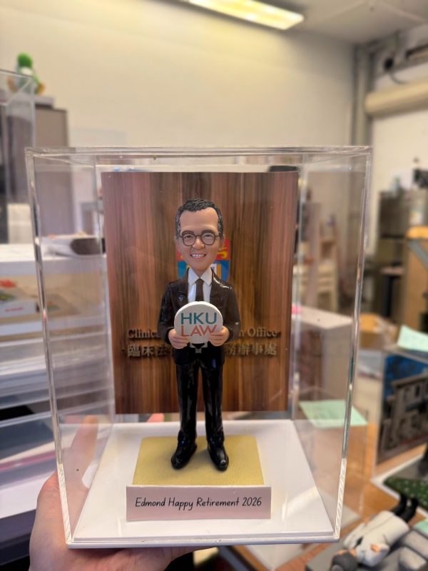 透明展示盒中的場景公仔：角色穿黑色西裝與領帶、戴圓框眼鏡，雙手捧圓形標牌，上有「HKU LAW」字樣；足下為淺金色方形底座。前方淺色銘牌寫有「Edmond Happy Retirement 2026」。背板為直紋木質背景並見英文字樣裝飾。