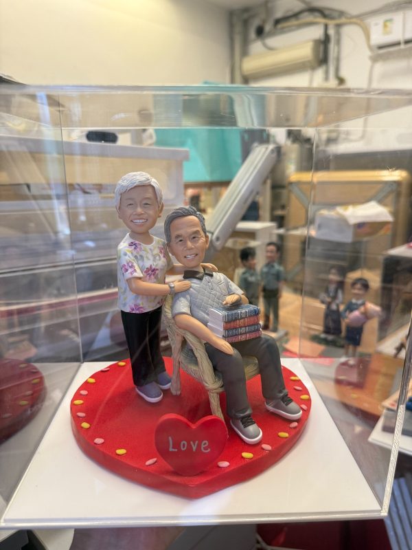透明展示盒中的雙人場景公仔：一位角色坐在淺色藤編椅上，懷抱成疊書本；另一位角色站在身後、手搭肩膀，穿碎花上衣與黑褲；坐者穿灰色上衣與深色長褲、運動鞋。兩人站坐於紅色愛心形底座，邊緣點綴彩色小圓片，前方有紅色愛心標牌寫著「Love」。
