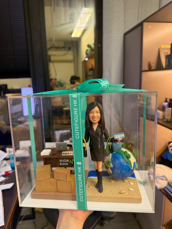 透明展示盒內的場景公仔：角色穿黑色套裝與長靴，手持藍色護照與登機證樣式卡片，另一手拎購物袋與藍色手提包；身後有藍白相間的地球造景與小型沙灘地面。左側是辦公桌（上放筆電、文件、咖啡杯）與一疊紙箱；桌前標示「NVDA $1000」字樣。展示盒外綁有綠松石色緞帶印有「CUTEFIGURE HK」。背景為室內辦公空間。