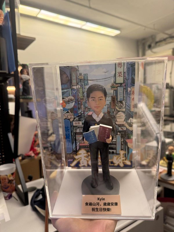 透明展示盒中的公仔：角色穿黑色西裝白襯衫，雙手各抱一本藍、棕色書本；腳下為灰色圓形底座。背板是色彩繽紛的城市街景與大型廣告牌。前方銅色小銘牌黑字寫有「Kyle」與中文生日祝福語。