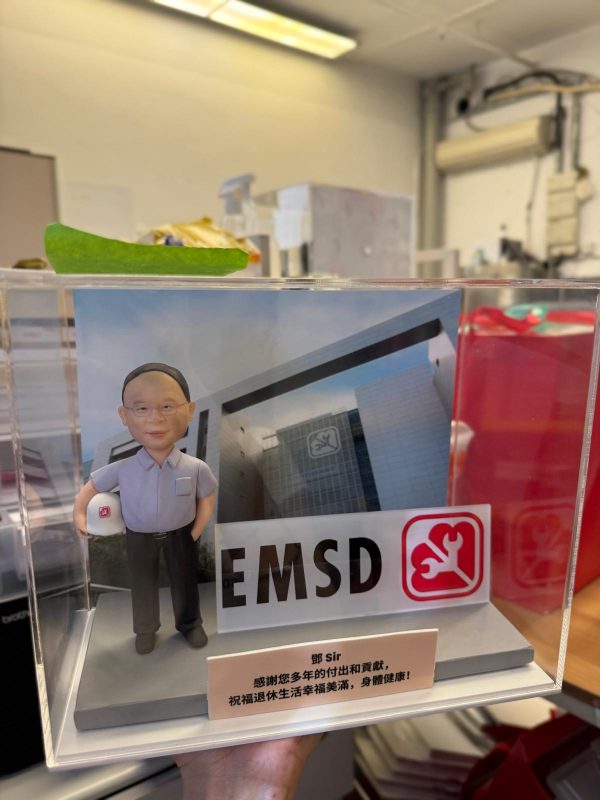 透明展示盒內的場景公仔：角色戴灰色短髮、穿淺灰襯衫與深色長褲、黑鞋，左手握一頂白色安全帽（側面印紅色扳手標誌）。背板為現代感玻璃外牆建築，上貼有同款圓角紅底白色工具標。前方立有大型「EMSD」字樣與紅色工具圖示的立體牌。底部灰色平台前有淺色銘牌，寫著繁體中文感謝賀詞與「鄺 Sir」稱呼。