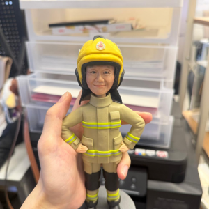 手持展示的消防主題公仔:角色戴黃色安全帽,帽前有圓形徽章;穿米色消防防護衣與褲,袖口與下擺、褲腳皆有螢光黃反光條;黑色長靴,雙手叉腰站在灰色圓形底座上。背景為室內層架與印表設備。