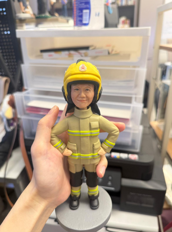 手持展示的消防主題公仔：角色戴黃色安全帽，帽前有圓形徽章；穿米色消防防護衣與褲，袖口與下擺、褲腳皆有螢光黃反光條；黑色長靴，雙手叉腰站在灰色圓形底座上。背景為室內層架與印表設備。