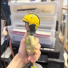 手持展示的消防主題公仔：角色戴黃色安全帽，帽前有圓形徽章；穿米色消防防護衣與褲，袖口與下擺、褲腳皆有螢光黃反光條；黑色長靴，雙手叉腰站在灰色圓形底座上。背景為室內層架與印表設備。