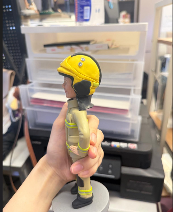 手持展示的消防主題公仔：角色戴黃色安全帽，帽前有圓形徽章；穿米色消防防護衣與褲，袖口與下擺、褲腳皆有螢光黃反光條；黑色長靴，雙手叉腰站在灰色圓形底座上。背景為室內層架與印表設備。