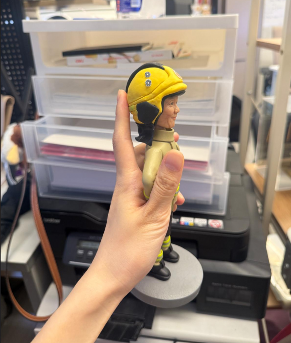 手持展示的消防主題公仔：角色戴黃色安全帽，帽前有圓形徽章；穿米色消防防護衣與褲，袖口與下擺、褲腳皆有螢光黃反光條；黑色長靴，雙手叉腰站在灰色圓形底座上。背景為室內層架與印表設備。