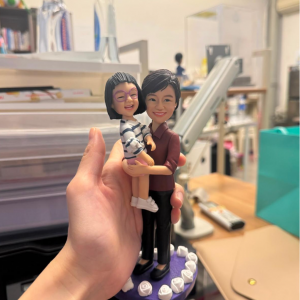 手持展示的親子主題公仔：成年角色穿酒紅上衣與黑長褲、黑色平底鞋，懷抱穿白底深色條紋洋裝與紫框眼鏡的小女孩；兩人面帶笑容。腳下為紫色多邊形底座，周圍環繞白色玫瑰裝飾。背景為室內桌面與收納物件。