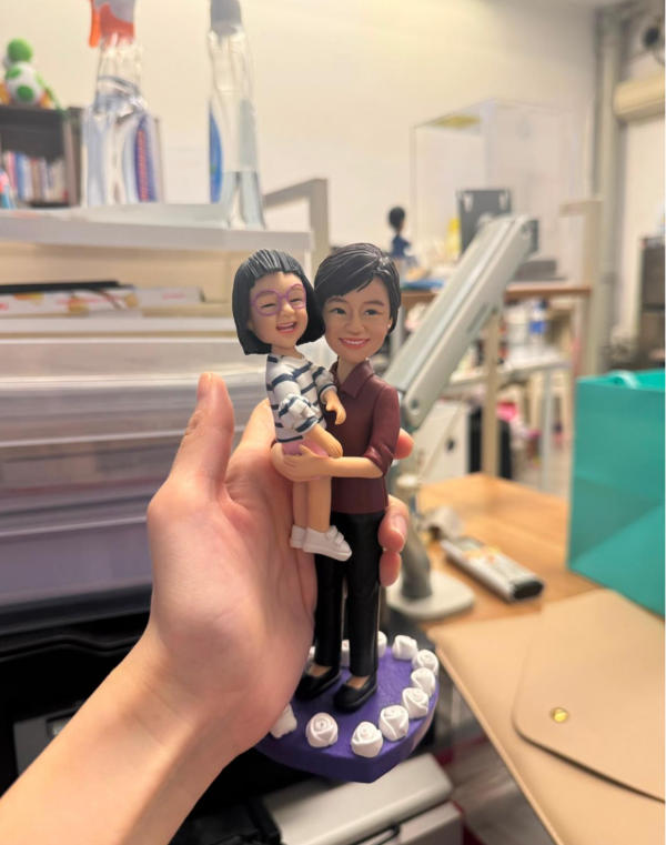手持展示的親子主題公仔：成年角色穿酒紅上衣與黑長褲、黑色平底鞋，懷抱穿白底深色條紋洋裝與紫框眼鏡的小女孩；兩人面帶笑容。腳下為紫色多邊形底座，周圍環繞白色玫瑰裝飾。背景為室內桌面與收納物件。