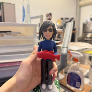 手持展示的休閒釣魚主題公仔:角色留中長髮戴黑色墨鏡,穿深藍拉鍊外套、深色長褲與白鞋,雙手抱著一條鮮紅色魚;腳下為灰色磚紋底座,兩側綠色草地點綴小花。背景為室內桌面與清潔噴瓶等物件。