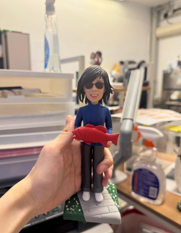 手持展示的休閒釣魚主題公仔：角色留中長髮戴黑色墨鏡，穿深藍拉鍊外套、深色長褲與白鞋，雙手抱著一條鮮紅色魚；腳下為灰色磚紋底座，兩側綠色草地點綴小花。背景為室內桌面與清潔噴瓶等物件。