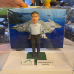 透明展示盒中的退休主題公仔：角色穿淡藍襯衫與深灰長褲，雙手放於身後站立；腳下為綠色草毯式矩形底座。背板為環海島嶼機場與多條跑道的鳥瞰圖。前方兩個小立牌：左側為綠色拱形圖案與「機場管理局 AIRPORT AUTHORITY HONG KONG」字樣；右側白底黑字為「Happy Retirement to Daniel Sir」。