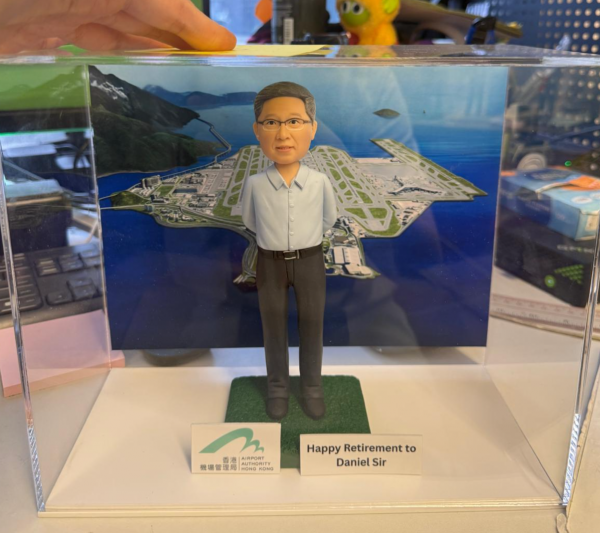 透明展示盒中的退休主題公仔：角色穿淡藍襯衫與深灰長褲，雙手放於身後站立；腳下為綠色草毯式矩形底座。背板為環海島嶼機場與多條跑道的鳥瞰圖。前方兩個小立牌：左側為綠色拱形圖案與「機場管理局 AIRPORT AUTHORITY HONG KONG」字樣；右側白底黑字為「Happy Retirement to Daniel Sir」。